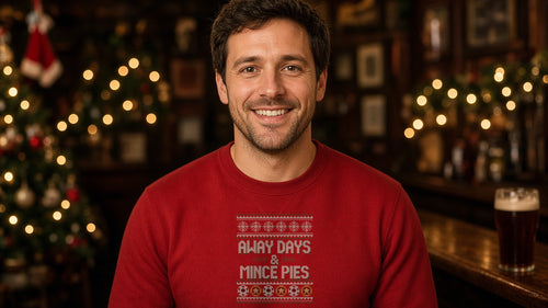 Man smiling in a cosy Christmas pub wearing the red Away Days & Mince Pies Christmas sweatshirt with a festive knit-pattern print